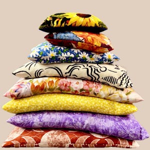 May include: A stack of eight decorative throw pillows with various floral and abstract patterns. The pillows feature a range of colors, including yellow, purple, blue, and red. The top pillow has a sunflower design. The pillows are stacked on top of each other.