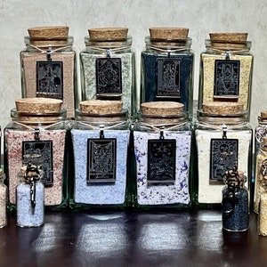 May include: A collection of glass jars filled with colorful salt and topped with cork lids. Each jar has a silver pendant with a black and white image of a tarot card. The jars are arranged in two rows on a wooden surface.