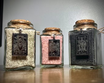 May include: Three glass jars with cork lids filled with different colored sand. Each jar has a silver charm hanging from the lid with a tarot card design. The charms read "The Chariot", "The Empress", and "The Emperor".