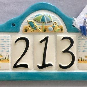 May include: A turquoise blue ceramic house number plaque with a beach scene featuring a beach umbrella, beach chairs, and a beach ball. The number 213 is displayed in black on white tiles.