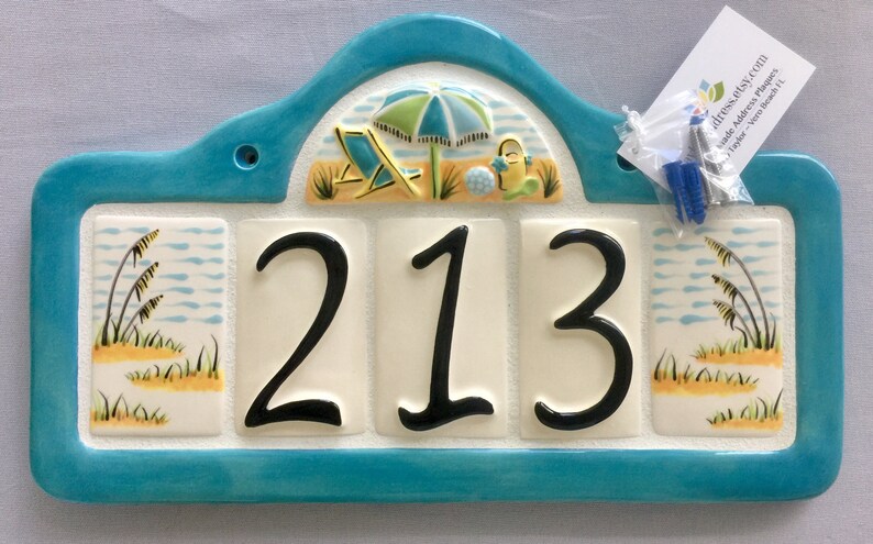 May include: A turquoise blue ceramic house number plaque with a beach scene featuring a beach umbrella, beach chairs, and a beach ball. The number 213 is displayed in black on white tiles.