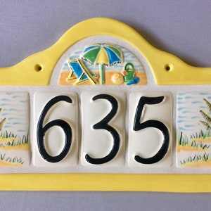 May include: Yellow ceramic house number sign with a beach scene and the number 635. The beach scene includes a beach umbrella, two beach chairs, a beach ball, and a bucket and shovel.