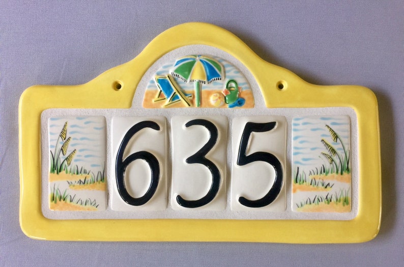 May include: Yellow ceramic house number sign with a beach scene and the number 635. The beach scene includes a beach umbrella, two beach chairs, a beach ball, and a bucket and shovel.