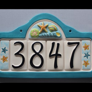 May include: A turquoise ceramic house number sign with a white background and black numbers. The numbers are 3847. The sign has a decorative top with a seashell, starfish, and a green and yellow flower.