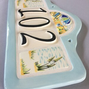 May include: A light blue ceramic house number sign with the number 201 in black lettering. The sign has a beach theme with images of palm trees, water, and an umbrella.