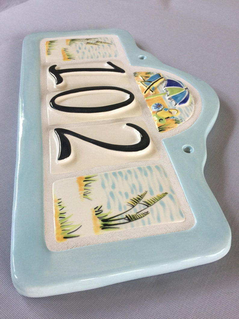 May include: A light blue ceramic house number sign with the number 201 in black lettering. The sign has a beach theme with images of palm trees, water, and an umbrella.