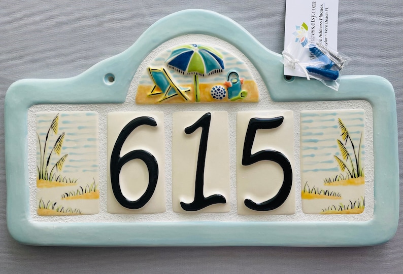 May include: A light blue ceramic house number sign with a beach theme. The number 615 is displayed in black on a white background. The top of the sign features a beach scene with an umbrella, beach chair, and a bucket and shovel. The sides of the sign feature a beach scene with tall grass.