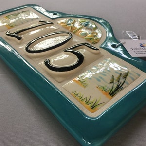 May include: A teal ceramic house number plaque with the number 105 in black lettering. The plaque has a decorative arch at the top and is decorated with images of palm trees, sand, and seagrass.