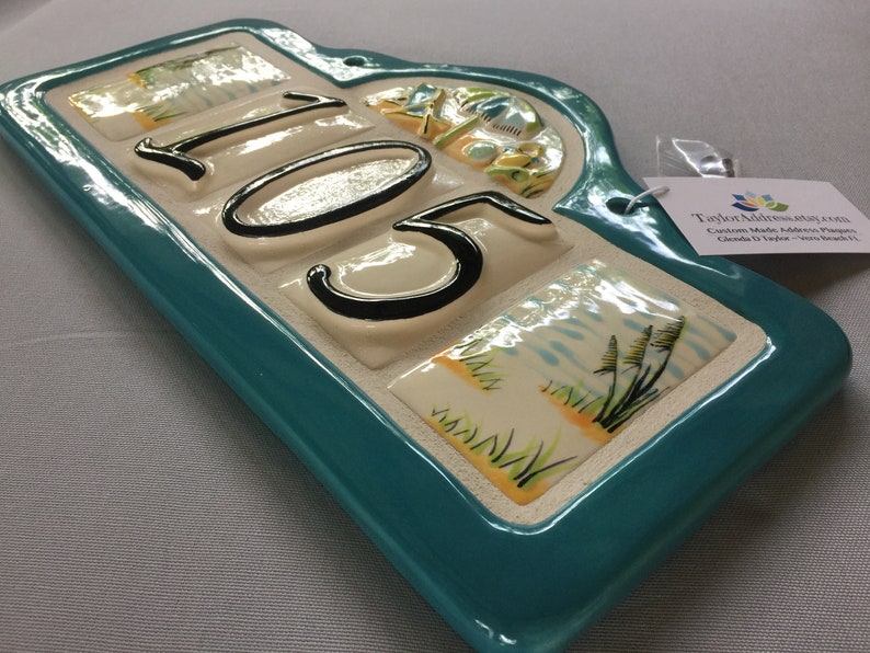May include: A teal ceramic house number plaque with the number 105 in black lettering. The plaque has a decorative arch at the top and is decorated with images of palm trees, sand, and seagrass.