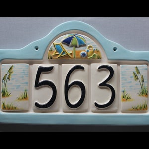 May include: A blue and white ceramic house number sign with a beach theme. The sign has a beach umbrella, beach chairs, and a watering can. The house number 563 is displayed in black on white tiles.