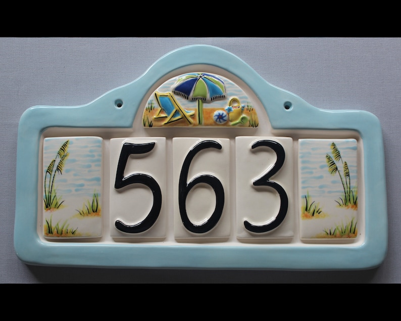 May include: A blue and white ceramic house number sign with a beach theme. The sign has a beach umbrella, beach chairs, and a watering can. The house number 563 is displayed in black on white tiles.