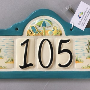 May include: A turquoise ceramic house number plaque with a beach scene featuring an umbrella, beach chair, and pail. The number 105 is displayed in black on a white background.