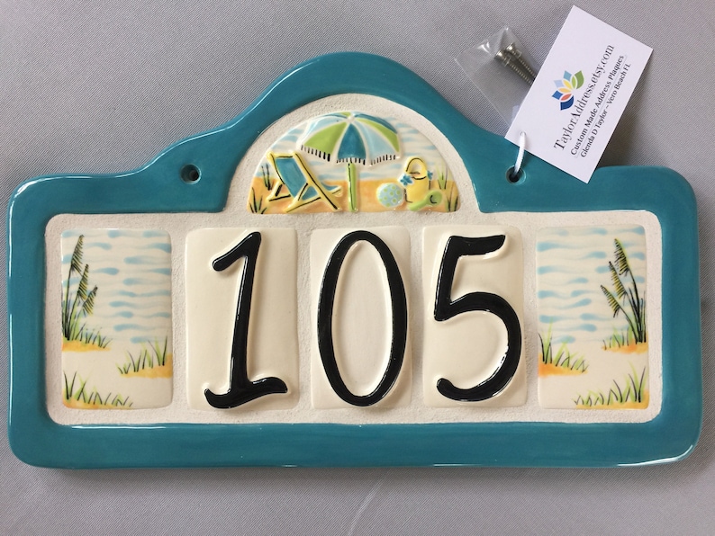 May include: A turquoise ceramic house number plaque with a beach scene featuring an umbrella, beach chair, and pail. The number 105 is displayed in black on a white background.