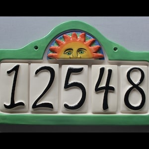 May include: Green ceramic house number sign with a sun face design and the numbers 12548.