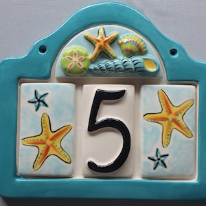 May include: A turquoise blue ceramic house number sign with a white background. The number "5" is in black with a blue outline. The sign is decorated with seashells, starfish, and sand dollars.