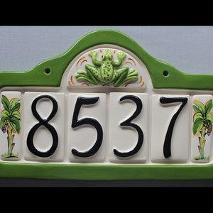 May include: Green ceramic address plaque with the numbers 8537 in black. The plaque has a green border and features a frog and palm tree designs. The background is a light blue.