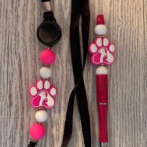 May include: A pink and white beaded retractable badge holder with a black lanyard and a pink pen with a paw print design. The pen has a white silhouette of a dog and cat.