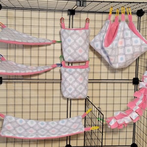 May include: A set of six pink and gray patterned hanging pet beds and a pink and white hanging toy for small animals. The beds are made of soft fabric and have a geometric pattern. The toy is made of soft fabric and has a knotted design.