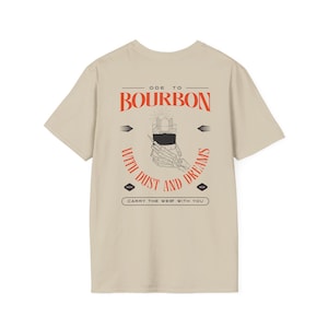 Könnte beinhalten: Beigefarbenes T-Shirt mit einem grafischen Druck einer Skeletthand, die ein Glas Bourbon hält. Der Text "Ode to Bourbon with Dust and Dreams" ist in Rot auf dem Shirt gedruckt. Der Text "Carry the West with You" ist in Schwarz unter dem Grafikdruck gedruckt.
