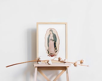 May include: A framed print of Our Lady of Guadalupe, a religious icon, in a brown frame. The print is on a wooden stool with a branch of dried cotton flowers.