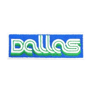 May include: Embroidered patch featuring the word "DALLAS" in white, outlined with green, on a blue rectangular background. The patch has a retro, sporty aesthetic, perfect for adding to clothing or accessories.