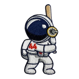 May include: An embroidered patch of an astronaut holding a baseball bat. The astronaut is wearing a white spacesuit with a blue helmet and the number 44 on the chest. The baseball bat is brown and has a yellow and black detail at the end.