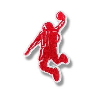 Könnte beinhalten: Ein roter, gestickter Aufnäher mit der Silhouette eines Basketballspielers beim Dunk. Der Aufnäher hat einen weißen Rand und ist vor einem weißen Hintergrund platziert. Der Aufnäher ist wahrscheinlich für Kleidung oder Accessoires.