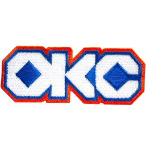 Puede incluir: Parche bordado con las letras "OKC" en blanco, delineadas en naranja y azul. Las letras tienen un diseño azul en forma de diamante en el centro. El parche está sobre un fondo blanco.