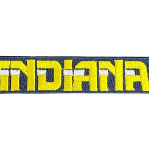 Puede incluir: Parche bordado con la palabra "INDIANA" en letras amarillas, contorneadas en blanco, sobre un fondo rectangular azul marino. El parche es rectangular y probablemente está diseñado para coserlo en ropa o accesorios.