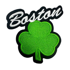 以下が含まれることがあります： 白い筆記体で「Boston」の文字が刺繍されたパッチ。緑色のクローバーの上に配置されています。パッチは黒い縁取りがあり、衣類やアクセサリーへの取り付けを想定したデザインです。
