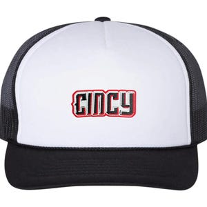 May include: A black and white trucker hat with a white front panel. The word "CINCY" is embroidered in red and black on the front. The hat has a black brim and a black mesh back.