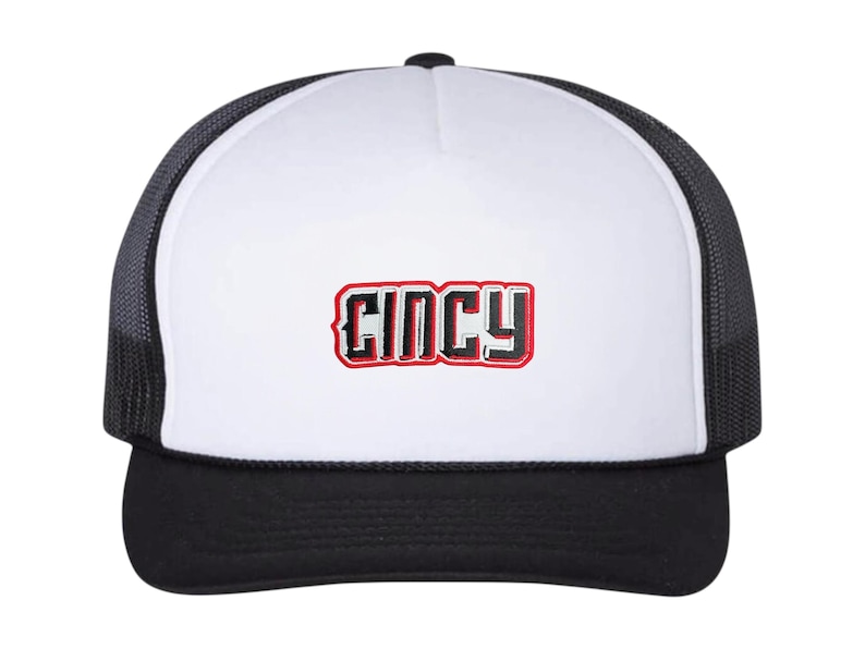 May include: A black and white trucker hat with a white front panel. The word "CINCY" is embroidered in red and black on the front. The hat has a black brim and a black mesh back.
