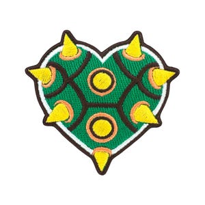 Peut inclure: Écusson brodé en forme de cœur, représentant une carapace de tortue verte avec des contours marron et des pointes jaunes. L'écusson a une bordure blanche et est destiné aux vêtements ou accessoires.