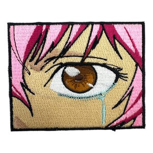 Könnte beinhalten: Gestickter Aufnäher mit einer Nahaufnahme eines Anime-Charakters mit einem einzelnen Tränen. Das Auge ist braun mit einer weißen Iris und einer blauen Träne. Der Charakter hat rosa Haare.