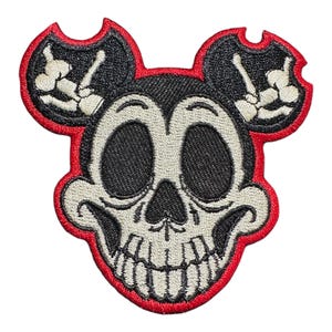 Puede incluir: Un parche bordado que representa una calavera con orejas de Mickey Mouse, en rojo, blanco y negro. La calavera sonríe y tiene una mano de esqueleto en cada oreja.