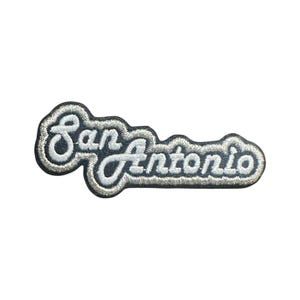 May include: Embroidered patch featuring the city name "San Antonio" in a cursive font. The patch has a white embroidered border and lettering, with a dark gray background.