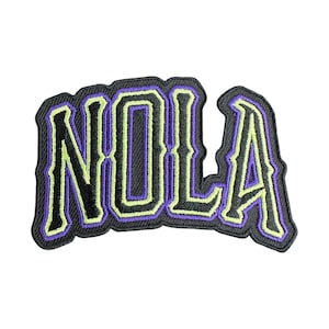 May include: Embroidered patch featuring the word "NOLA" in a stylized font. The letters are black with a purple outline and a light green inner border. The patch has a black border and is likely designed to be ironed or sewn onto fabric.