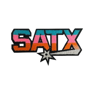 May include: Embroidered patch featuring the text "SATX" in bold, block letters. The letters are colored in a gradient of pink, orange, and teal, outlined in black. A starburst design is below the text, with a white and silver color scheme.