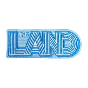 May include: A light blue embroidered patch featuring the words "THE LAND" in a retro style. The patch is a rounded rectangle with a light blue border and a white outline.