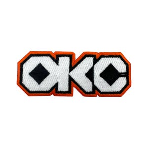 May include: Embroidered patch featuring the letters "OKC" in white, outlined in black, and bordered by orange. The letters have a geometric design with black diamond shapes. The patch is rectangular and likely for clothing or accessories.