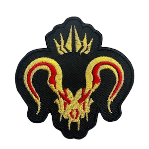 Op de afbeelding: Zwart geborduurde patch met een gestileerd ram schedel ontwerp met rode en gouden accenten. De patch heeft een unieke vorm en is waarschijnlijk voor kleding of accessoires.