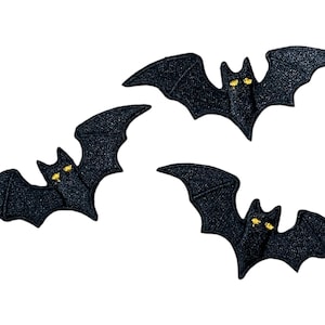 May include: Three black bat-shaped appliques with glittery texture and yellow eyes are displayed against a white background. These decorative patches are ideal for Halloween or craft projects.