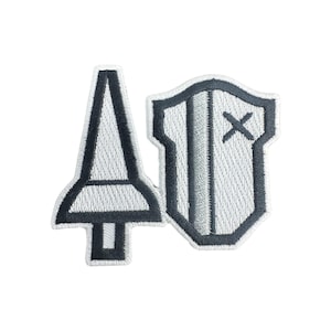 Könnte beinhalten: Gesticktes Patch-Set mit einem stilisierten Speer- und Schild-Design. Der Speer ist hellgrau mit schwarzer Umrandung, und der Schild ist ebenfalls hellgrau mit schwarzer Umrandung und einem 'X'-Detail. Die Patches sind wahrscheinlich für Kleidung oder Accessoires.