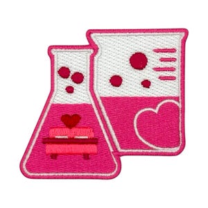 Puede incluir: Dos vasos de precipitados bordados de color rosa con corazones y fondo blanco. Los vasos de precipitados están llenos de un líquido rosa.