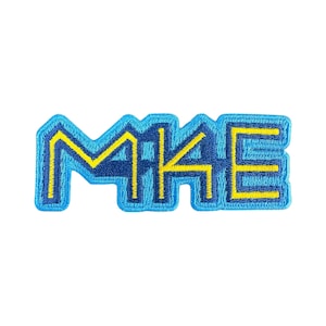 Könnte beinhalten: Gestickter Aufnäher in rechteckiger Form mit dem Wort "MAKE" in Großbuchstaben. Die Buchstaben sind blau mit gelben Umrissen und gelbem Innenraum. Der Aufnäher hat einen blauen Rand und ist vor einem weißen Hintergrund platziert.