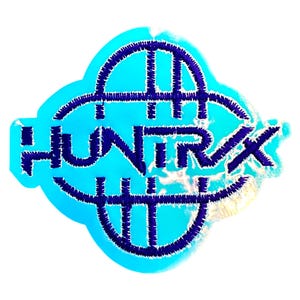 May include: A blue and white sticker with the word "HUNTRX" in a stylized font. The text is in dark blue, with a stitched appearance, set against a light blue background. The sticker has a unique, rounded shape.