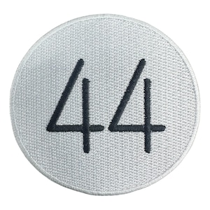 May include: A circular, embroidered patch with a light gray background. The number "44" is prominently displayed in black, stylized numerals. The patch has a textured appearance.