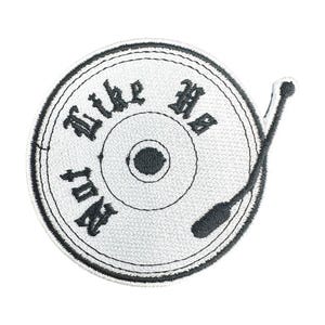 Op de afbeelding: Een witte geborduurde strijk-op patch met een zwarte vinylplaat en een naald. De plaat heeft de tekst "Not Like Us" rond de rand.