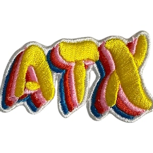 Puede incluir: Parche bordado con las letras "ATX" en un estilo retro, multicolor y 3D. Las letras son amarillas con contornos rosas, rojos y azules.