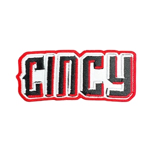 Op de afbeelding: Geborduurde patch met het woord "Cincy" in vette blokletters. De letters zijn zwart met een witte rand en een rode rand. De patch is rechthoekig en heeft een witte achtergrond.